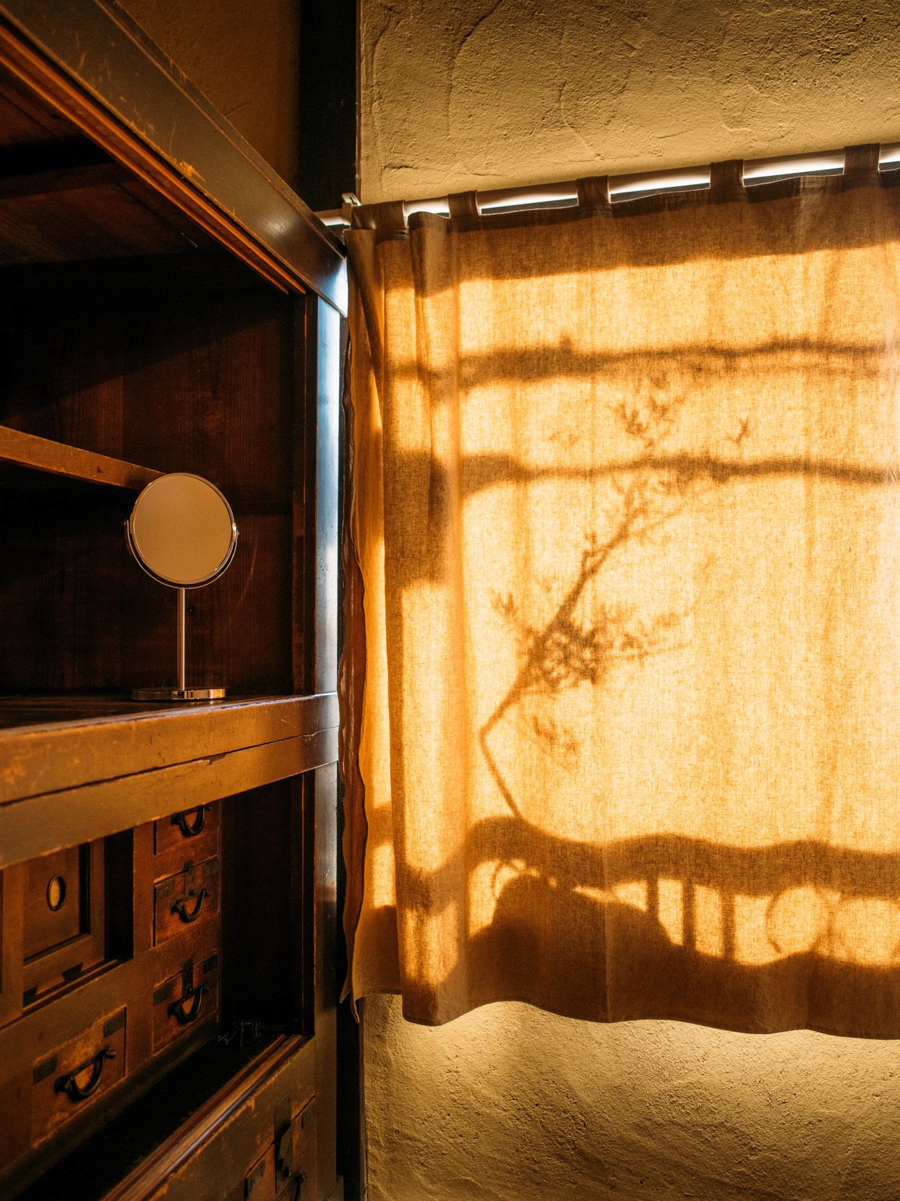 Sunlit wooden interior with mirror and drawers casting shadows on linen curtains at A Day in Khaki guesthouse in Kyoto.