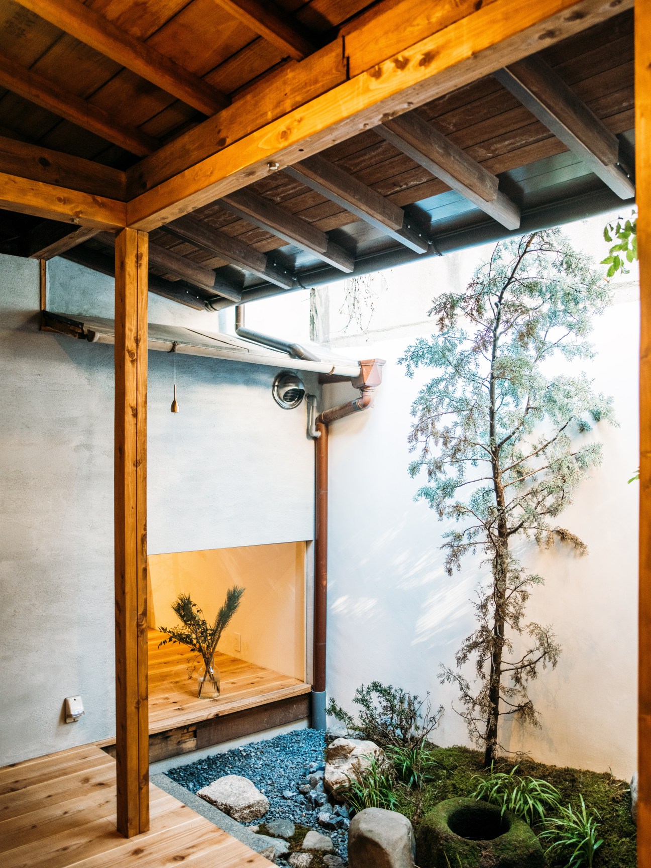Wooden pergola with beams overlooking a courtyard garden with gravel, stones, plants and a tall tree.