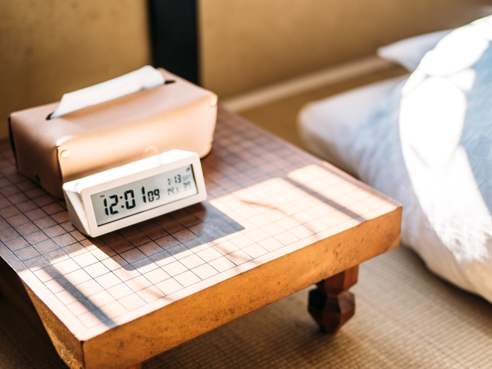 Digital clock displaying 12:00 on a gridded paper surface next to a tan leather tissue box on a wooden shelf.