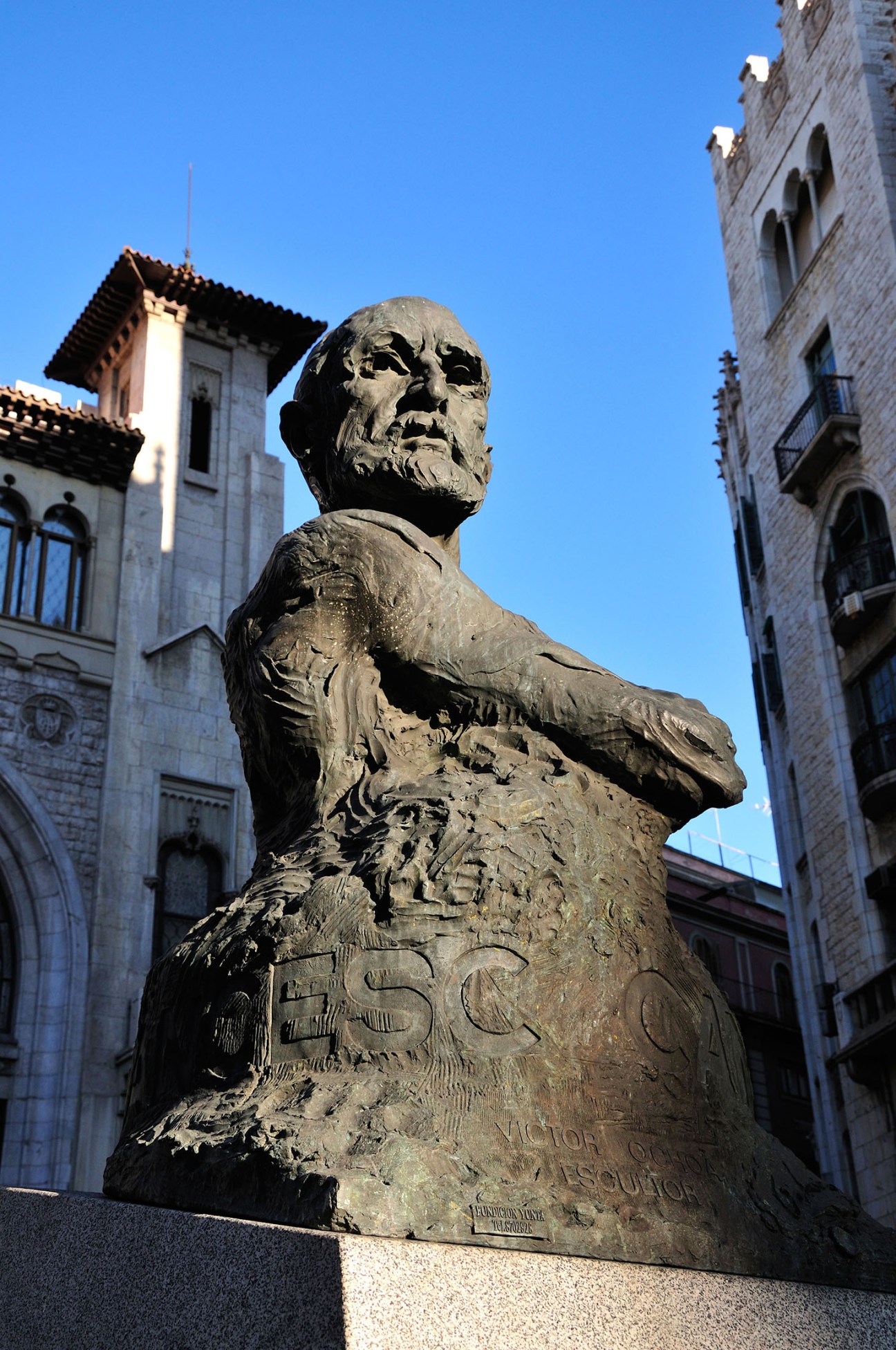 Stone bust statue of Francesc Cambó, Catalan independence champion, sculpted by Victor Ochea, flanked by historic buildings.