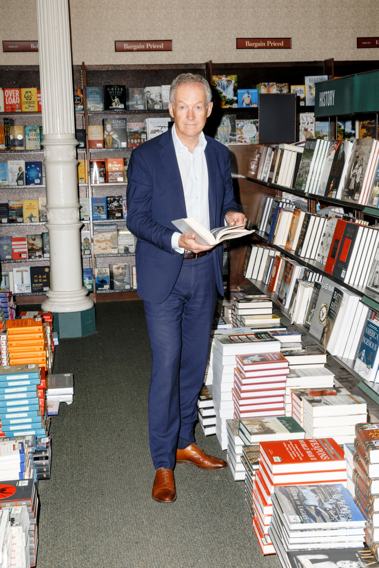 Man in dark suit holding open book while standing in bookstore aisle surrounded by shelves and stacks of books