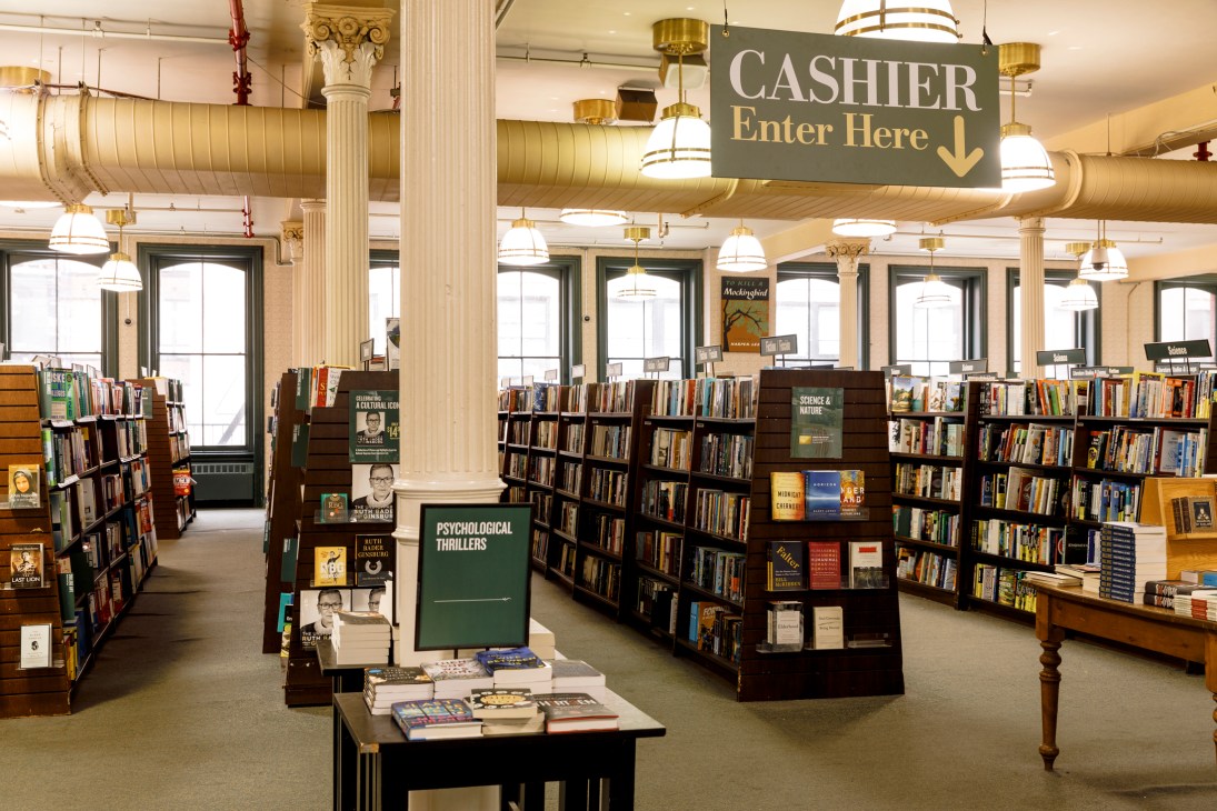 Interior of Barnes & Noble bookstore showing angled bookshelves, hanging sign reading CASHIER Enter Here, and pendant lighting