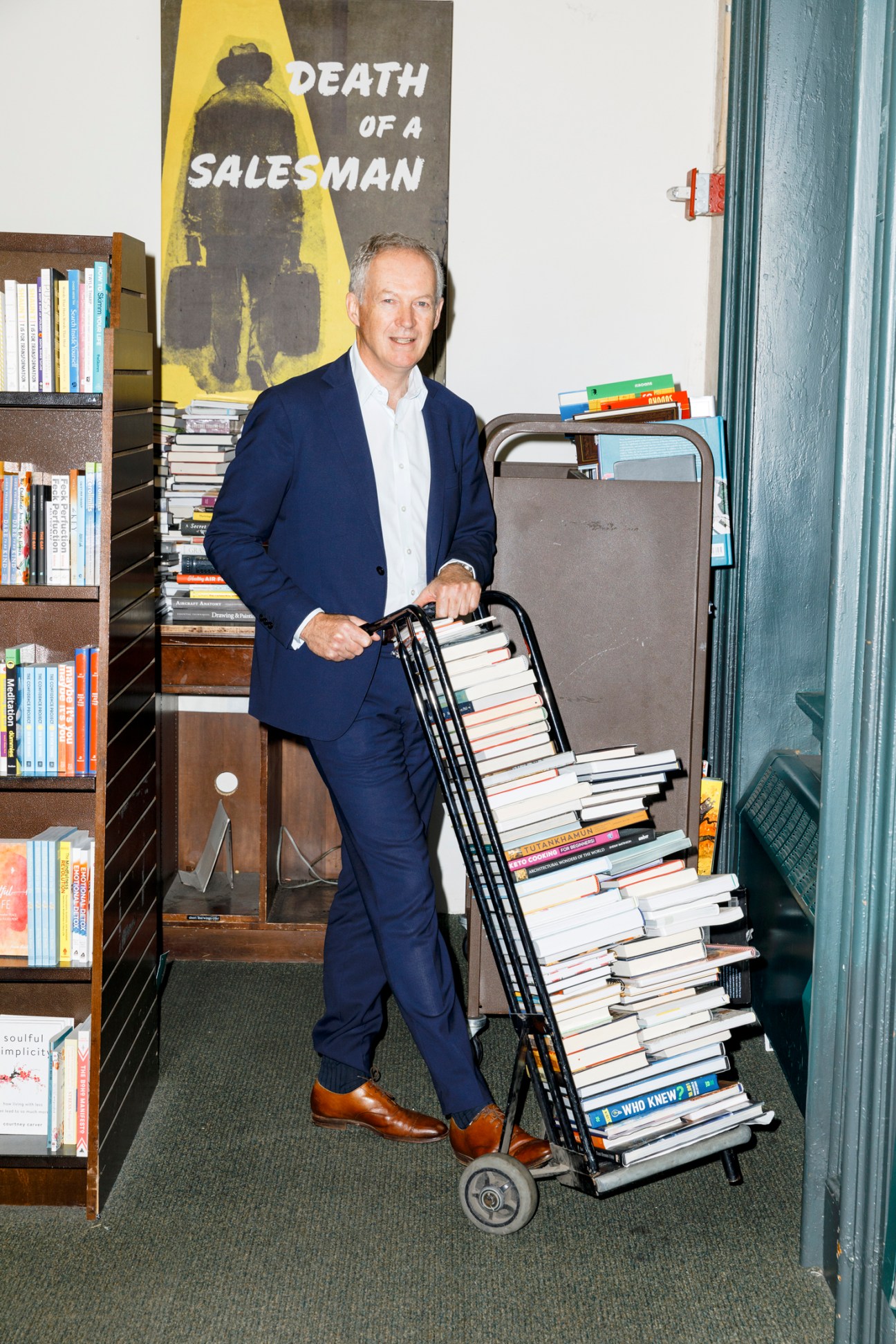 Man in navy suit pushing cart loaded with stacks of books in bookstore with shelves and 'Death of a Salesman' poster