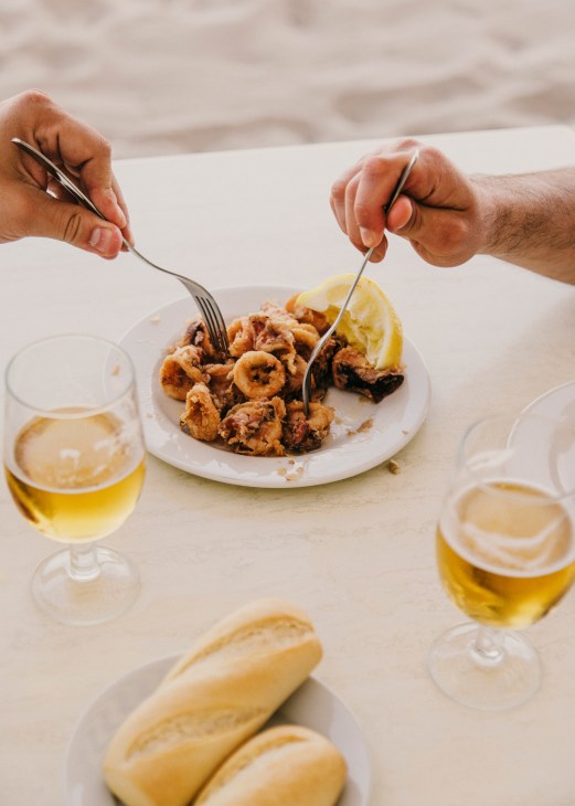 Two hands eating fried calamares from a white plate with lemon, white wine glasses nearby on a beach table.