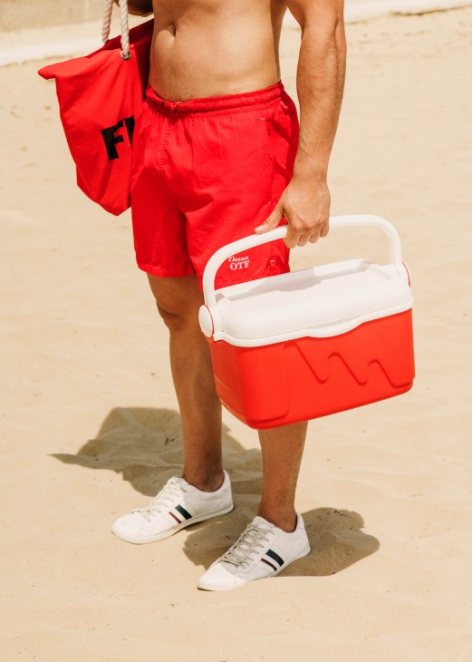 Man in red swim shorts holding a red and white cooler on a beach.