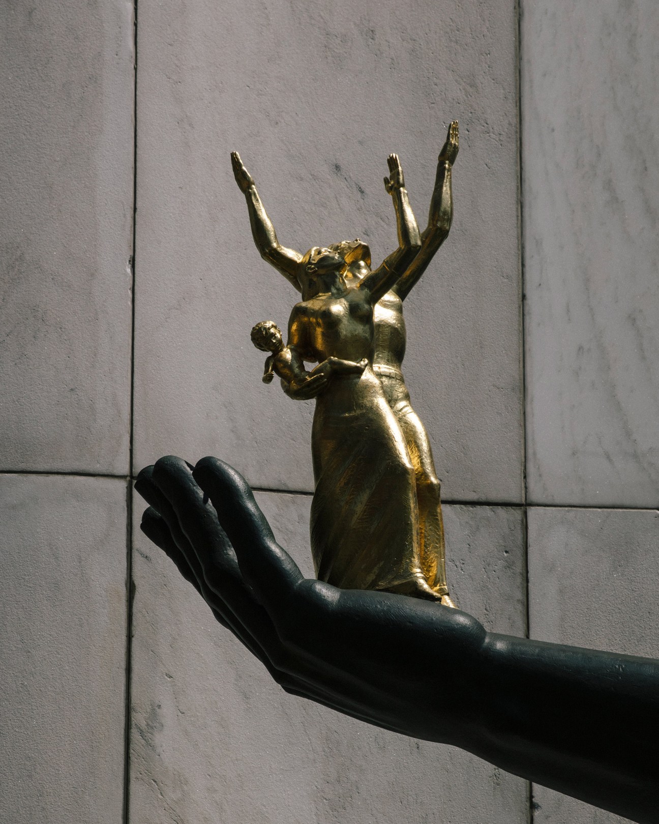 Close-up of a golden sculpture of a woman and child held in a large black hand against a white stone wall.