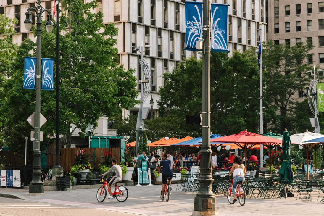 Hart Plaza in downtown Detroit with colorful umbrellas, outdoor seating, cyclists, and people enjoying the public space surrounded by tall buildings.