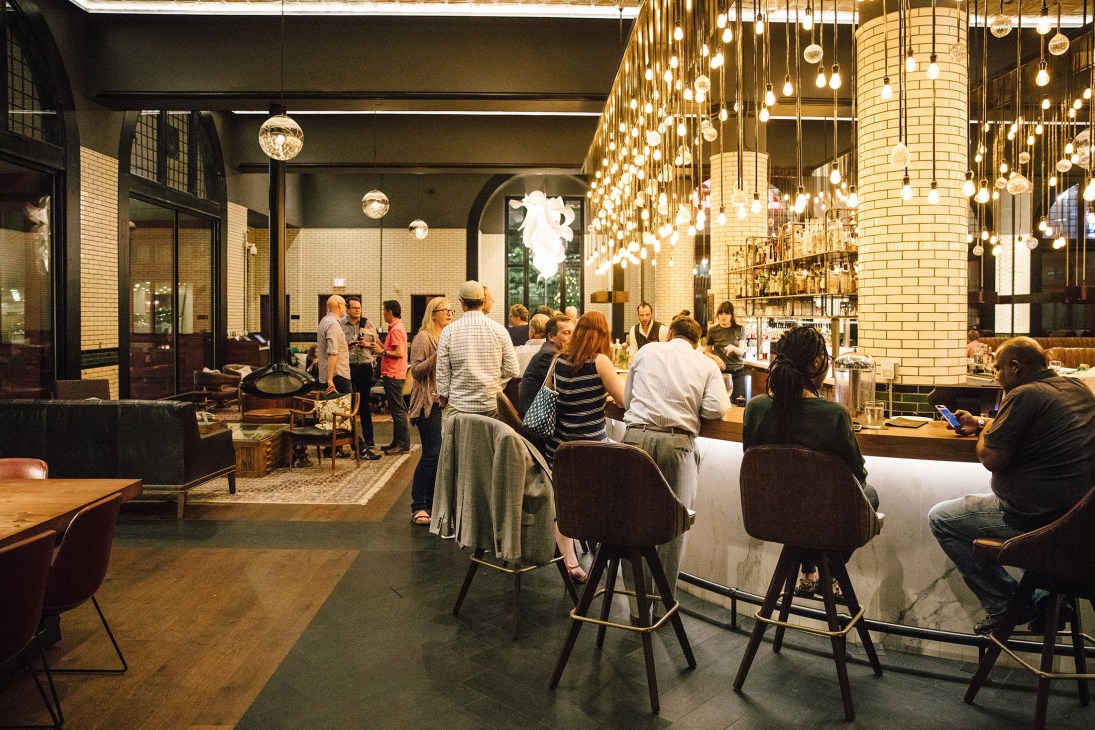 Interior of Foundation Hotel in Detroit Fire Department HQ with pendant lighting, brick columns, and patrons at bar and seating areas.