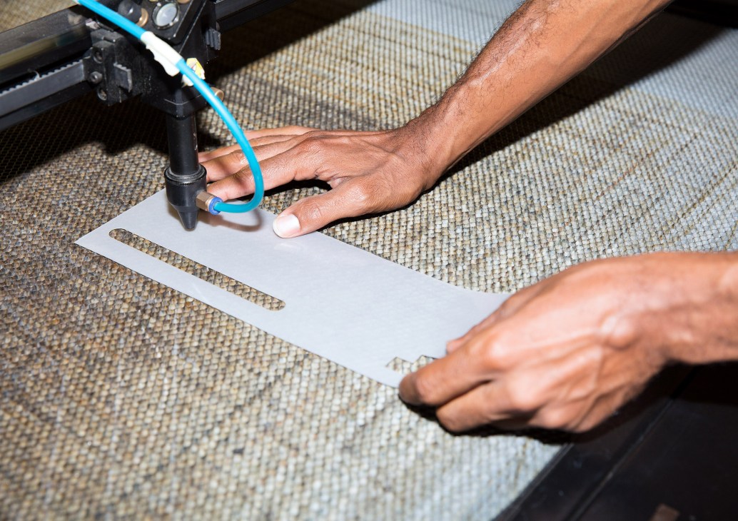 Hands operating a laser-cutting machine to cut white material from textured fabric.