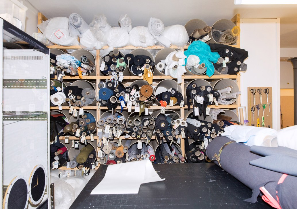 Wooden shelving unit filled with rolls of fabric samples, spools of thread, and textile materials in various colors organized on labeled spindles.