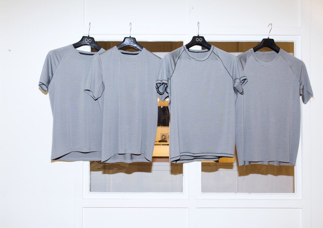 Four grey performance t-shirts hanging on a wooden rail with black hangers against a white wall.