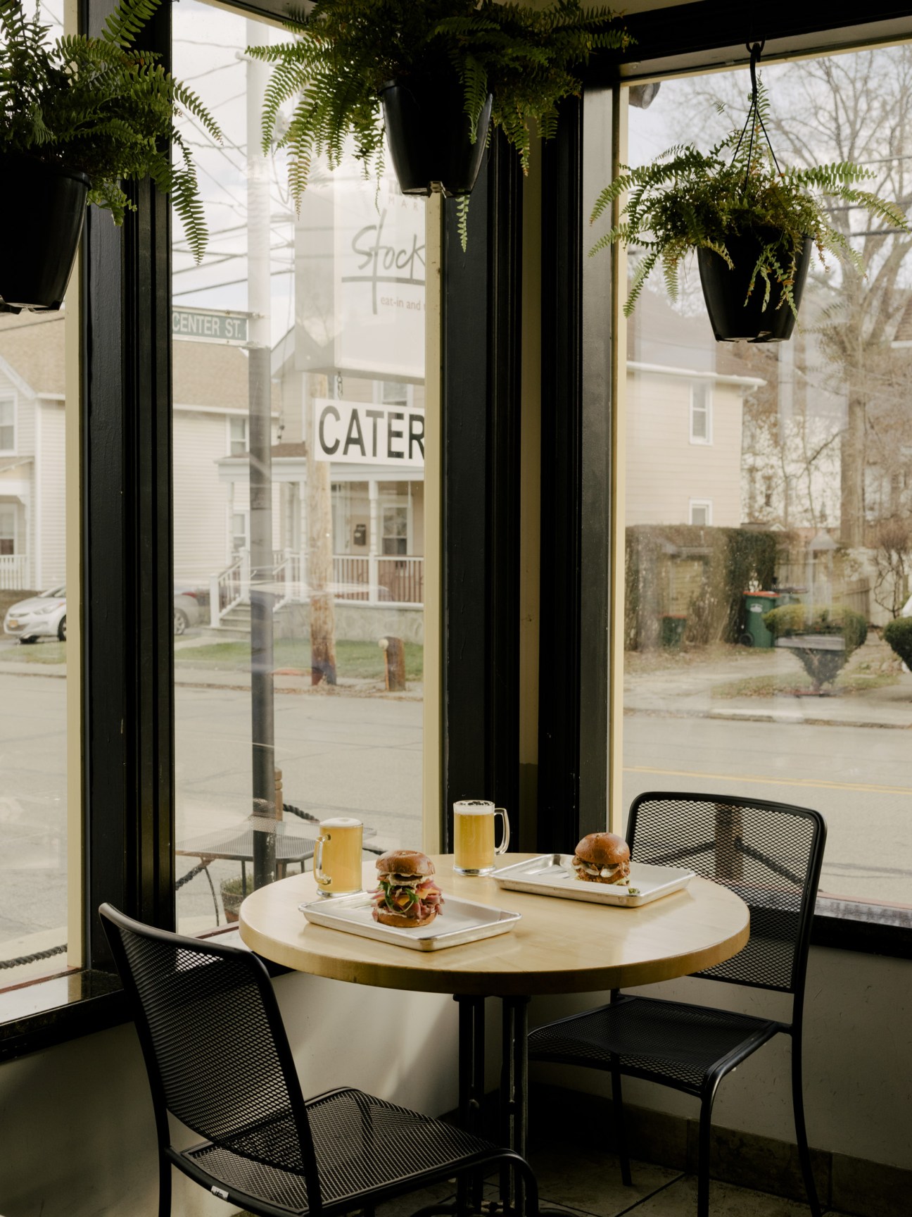 Interior of Fika bistro in Beacon with ferns hanging overhead, round table with burgers and beer beside large windows overlooking Main Street.