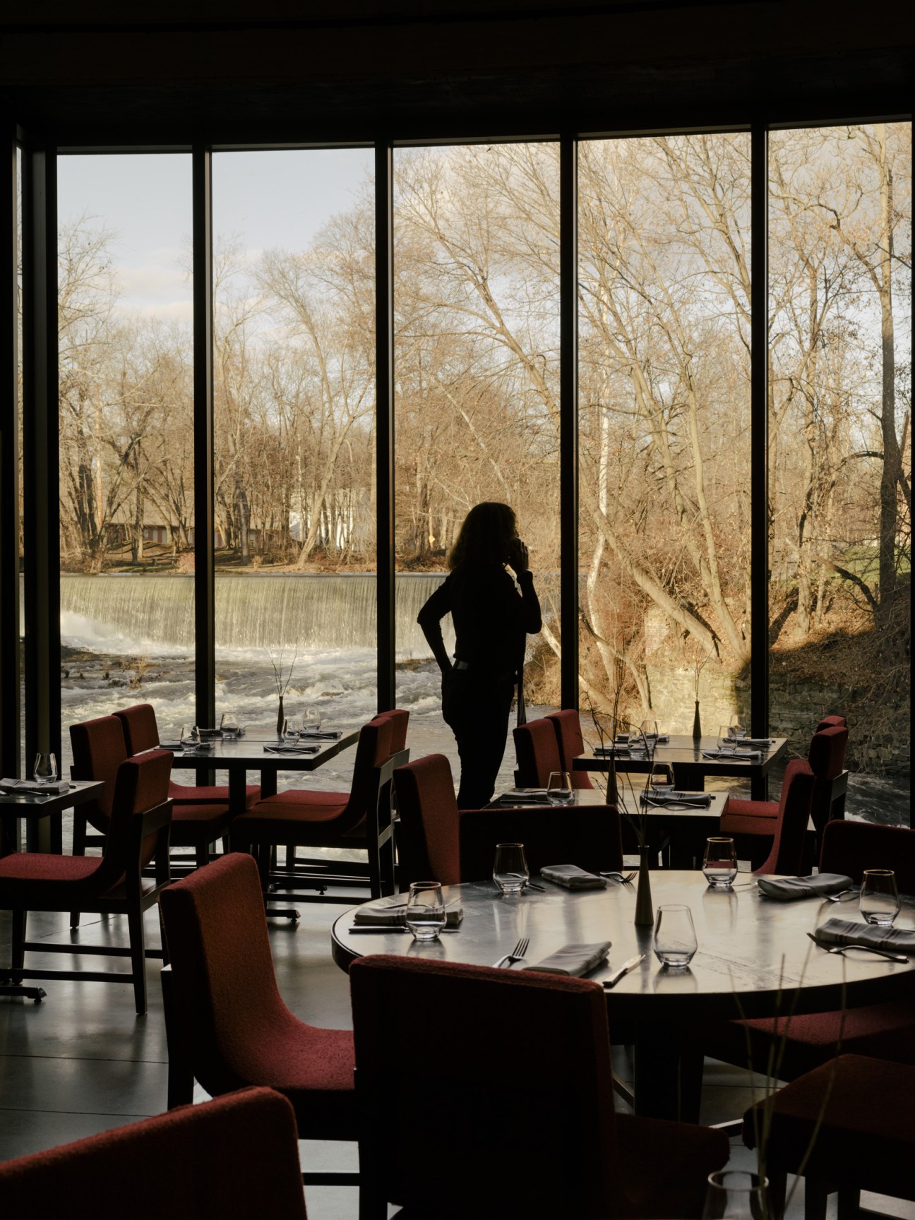 Woman standing in modern dining room overlooking bare trees and river through large windows.