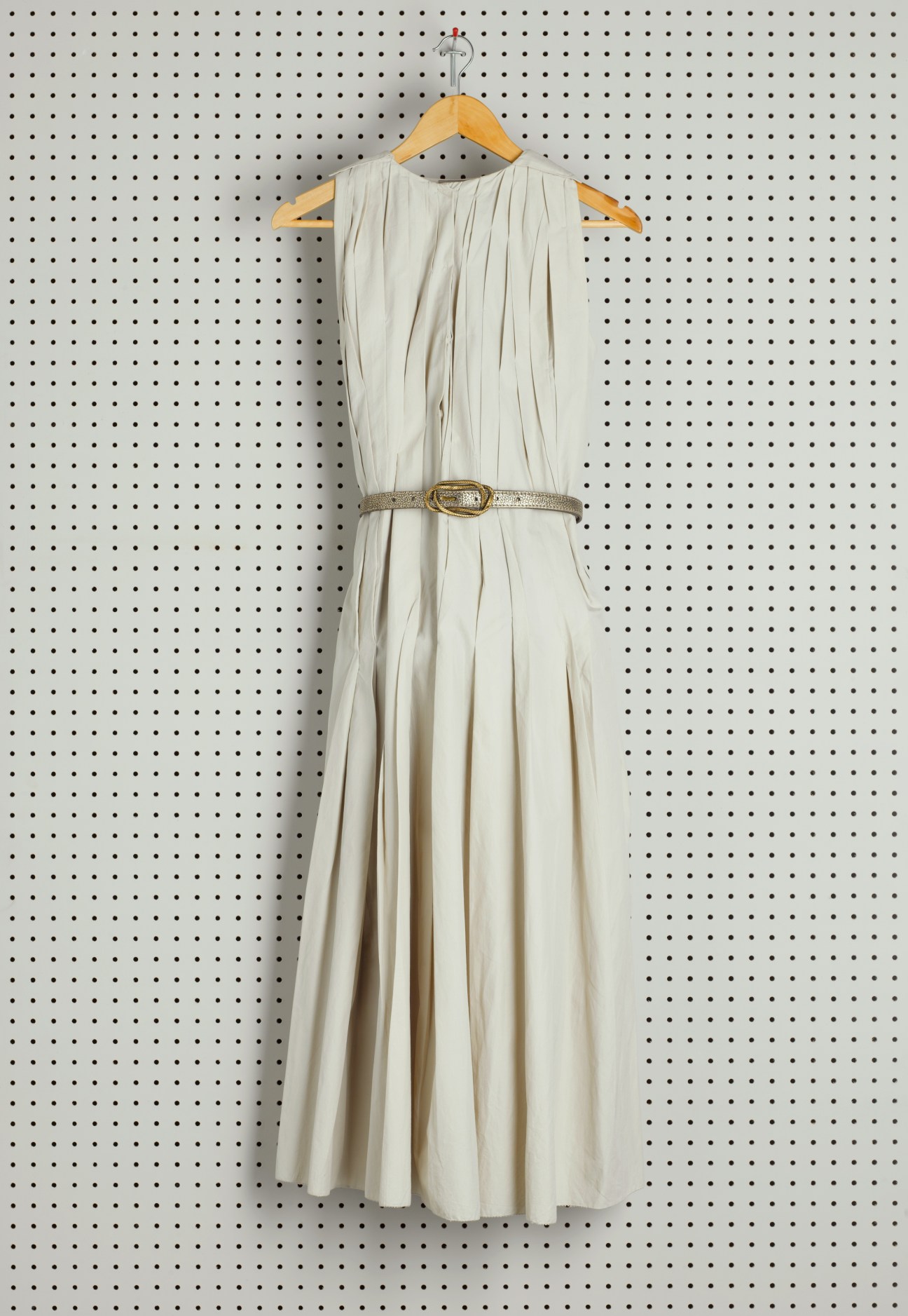 Cream-colored pleated sleeveless dress with gold belt on wooden hanger against pegboard wall.