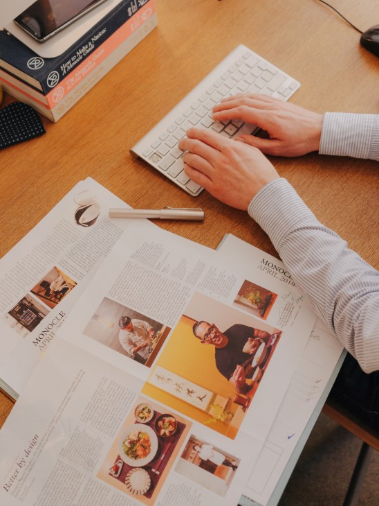 Person's hands typing on a keyboard while reviewing a Monocle magazine layout on a wooden desk.