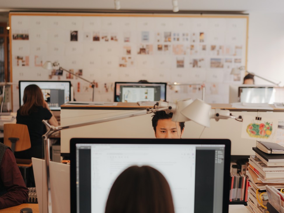 Monocle staff work at desks in an open office with mood boards and layouts pinned on the wall behind them.
