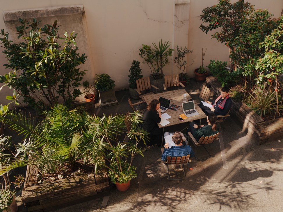 Monocle staff working at a wooden table in a sunny courtyard surrounded by potted plants.