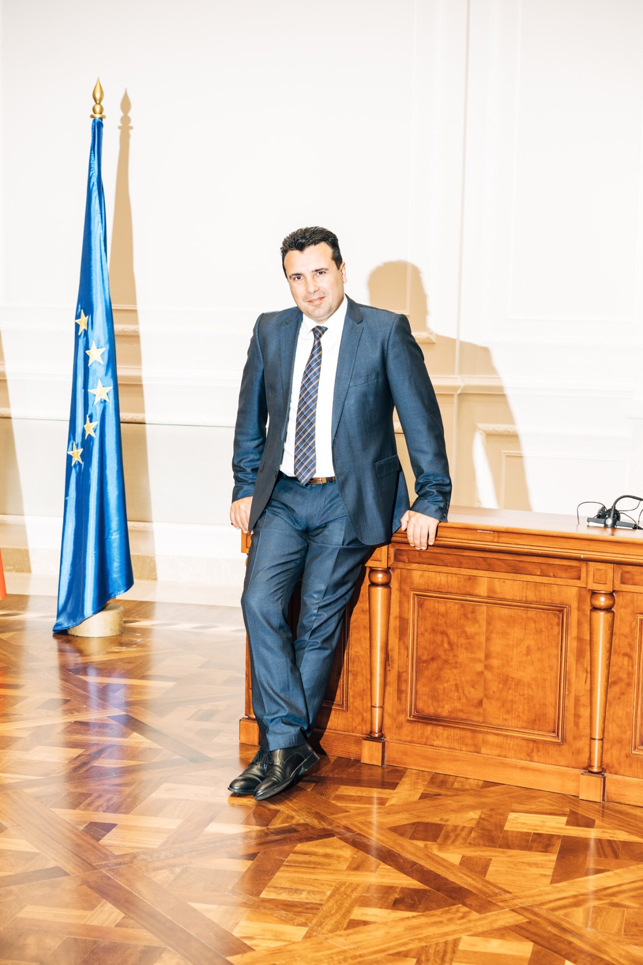 Zoran Zaev in a blue suit standing in an official government setting with EU flag.