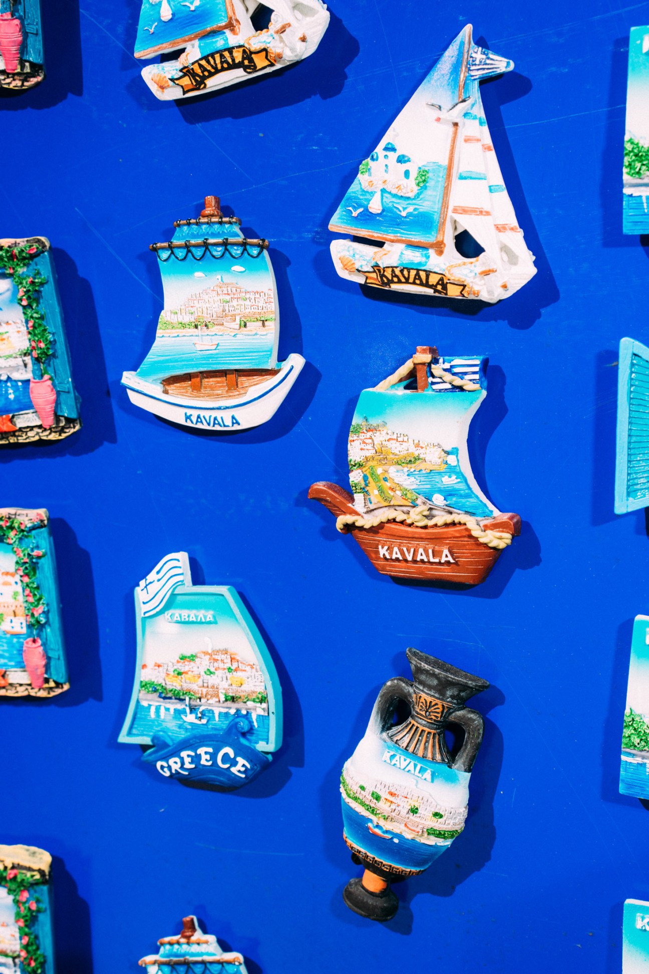 Colorful Kavala boat-shaped souvenirs and Greek magnets displayed on a bright blue surface.