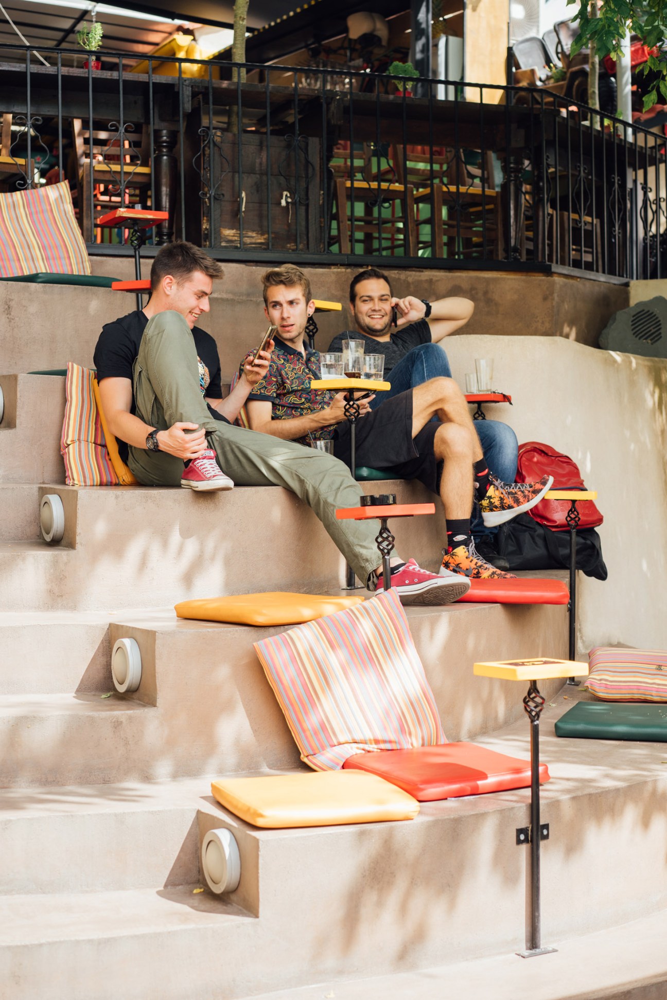 Three men sit on tiered concrete steps at Karaoli Square in Kavala, enjoying drinks and conversation.