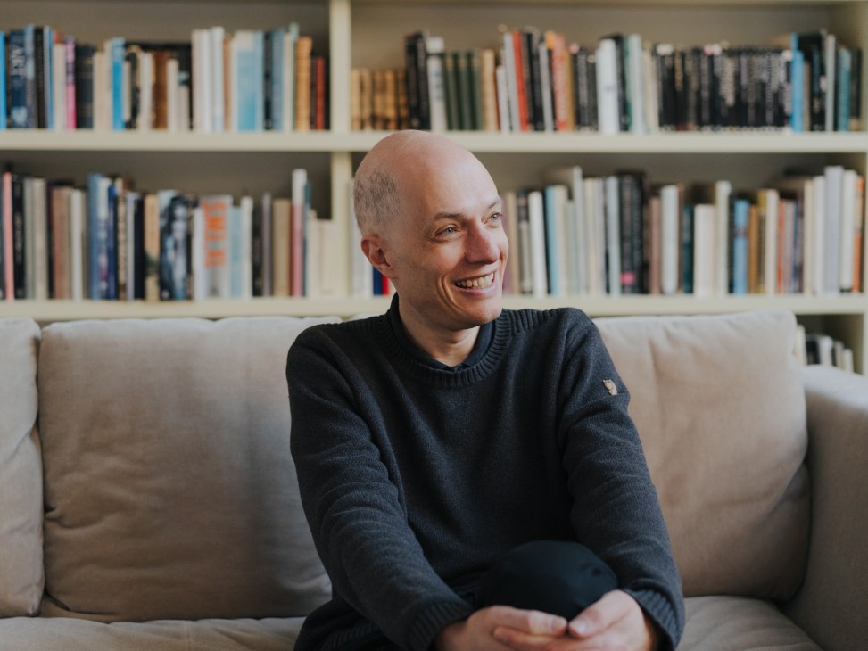 Alain de Botton seated on a beige sofa, smiling, with bookshelves behind him in his London home.
