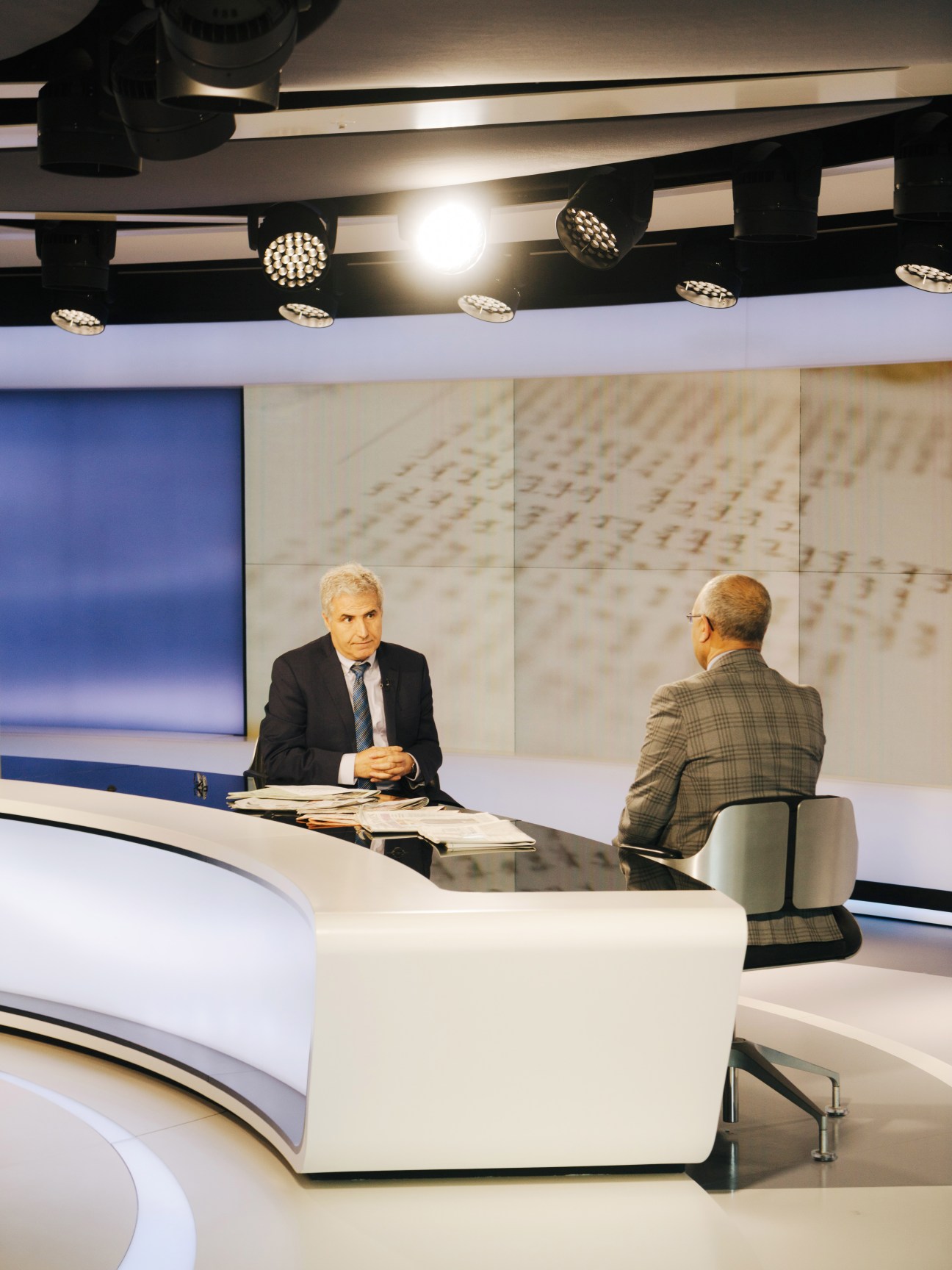 Two men conduct an interview at a curved desk in a modern television studio with blue screens and professional lighting.