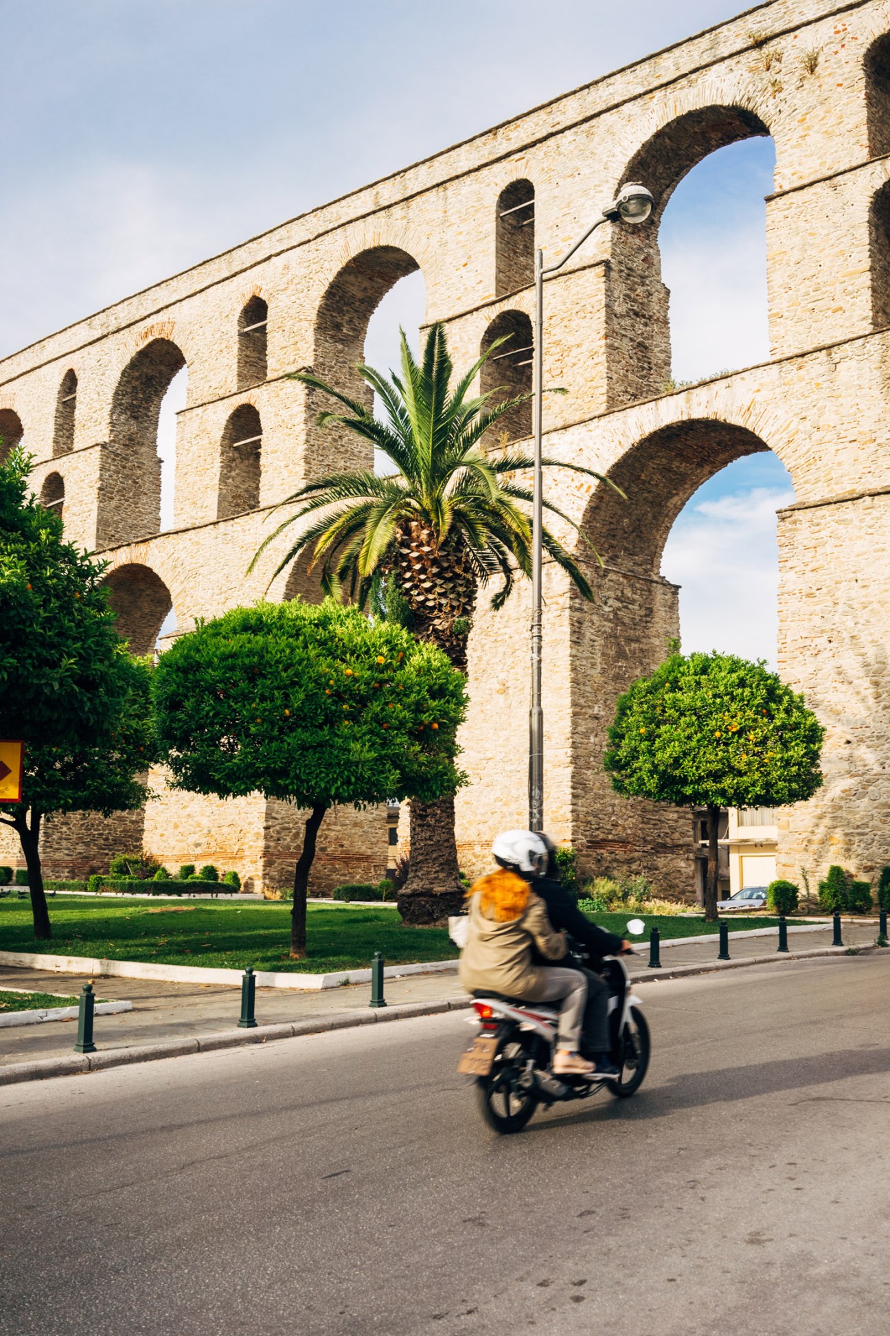Motorcyclist riding past the historic Kavala Aqueduct with arched stonework and palm tree.