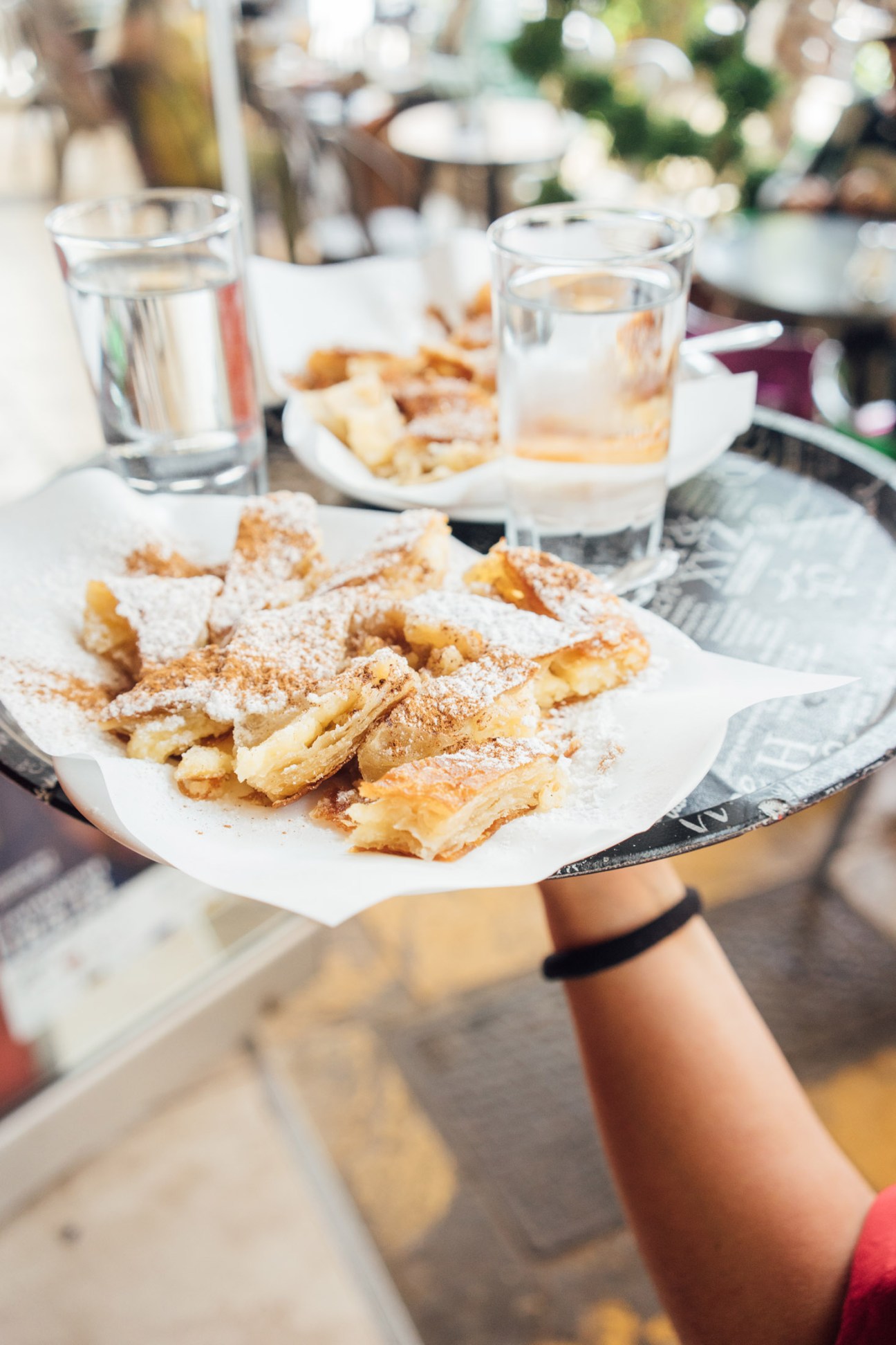 Hand holding a white plate of bougatsa pastries sprinkled with cinnamon and sugar at an outdoor café.
