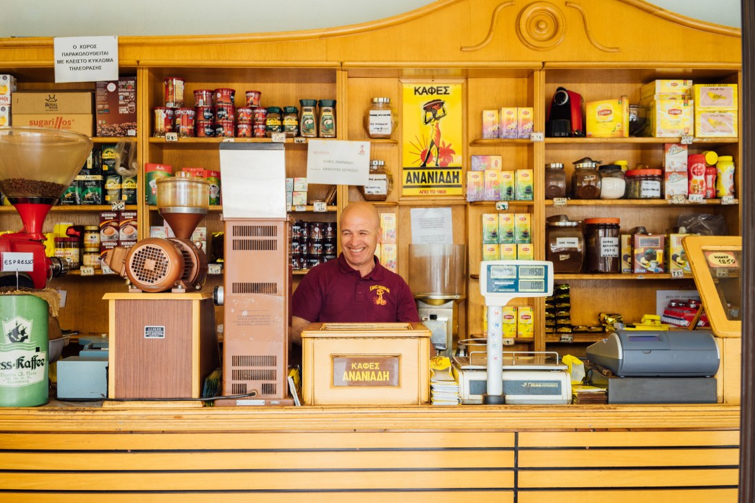 Owner Themistocles Ananiadis stands behind the counter of his family coffee roastery with shelves of coffee products and equipment.