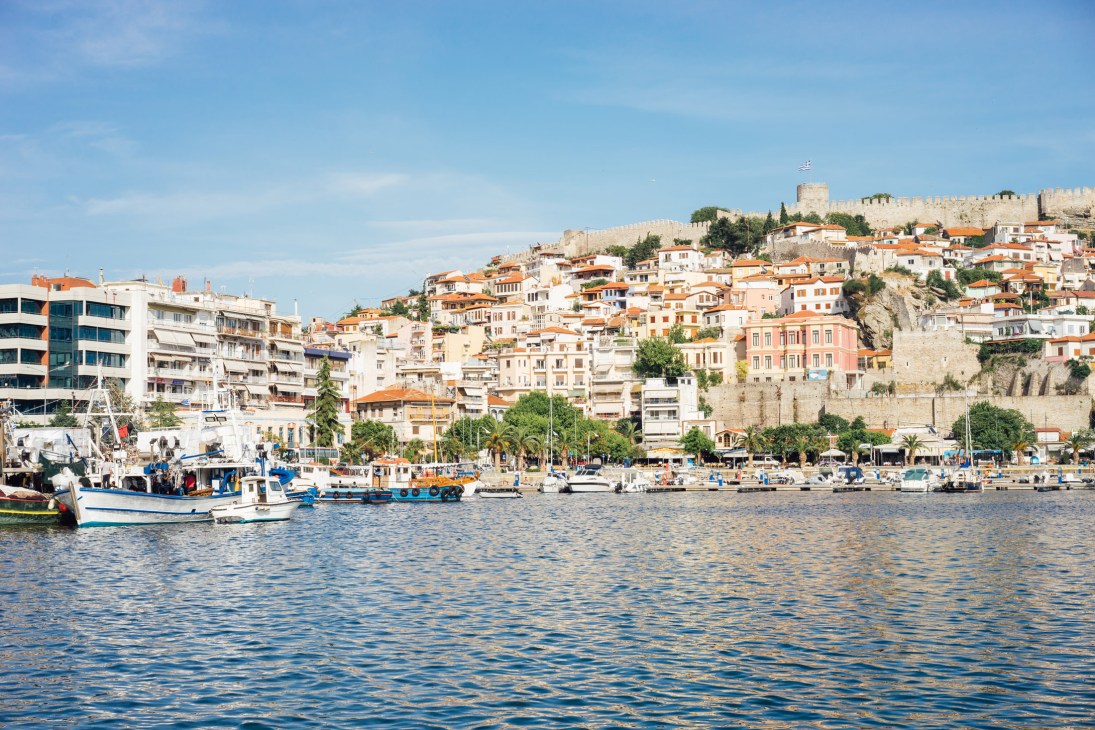 Kavala's harbourside old town with colorful buildings and ancient castle rising above, fishing boats moored in the foreground.