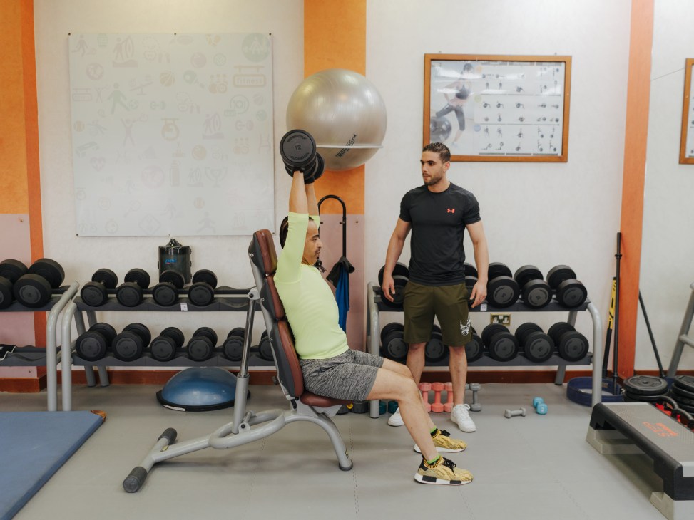 Woman lifting dumbbell on exercise equipment while man watches in gym.