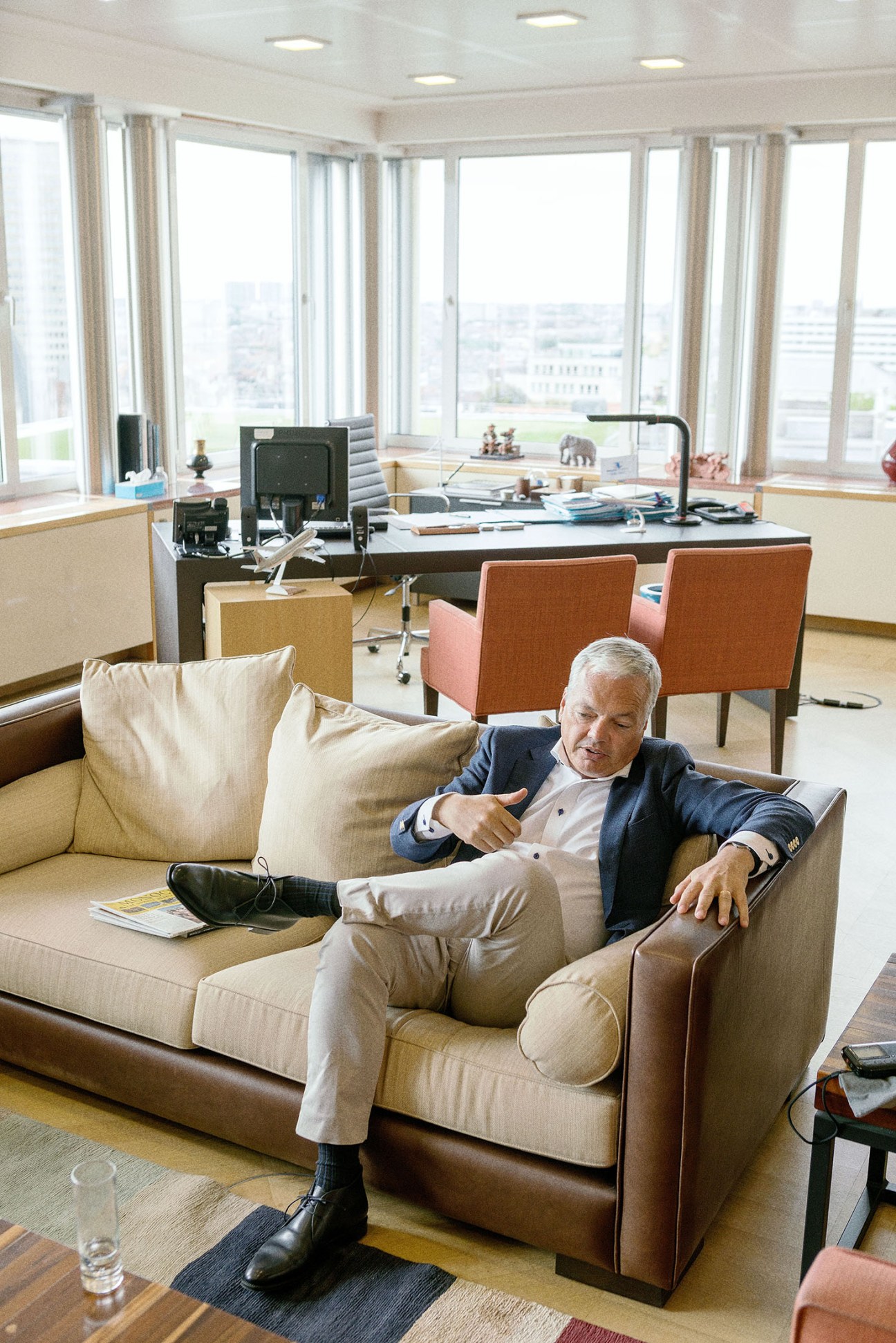 Belgian foreign minister Didier Reynders sitting on a beige sofa in a modern office with floor-to-ceiling windows overlooking a city.
