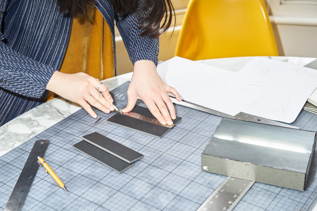 Designer's hands arranging architectural model materials on a grid-patterned work surface in a studio.