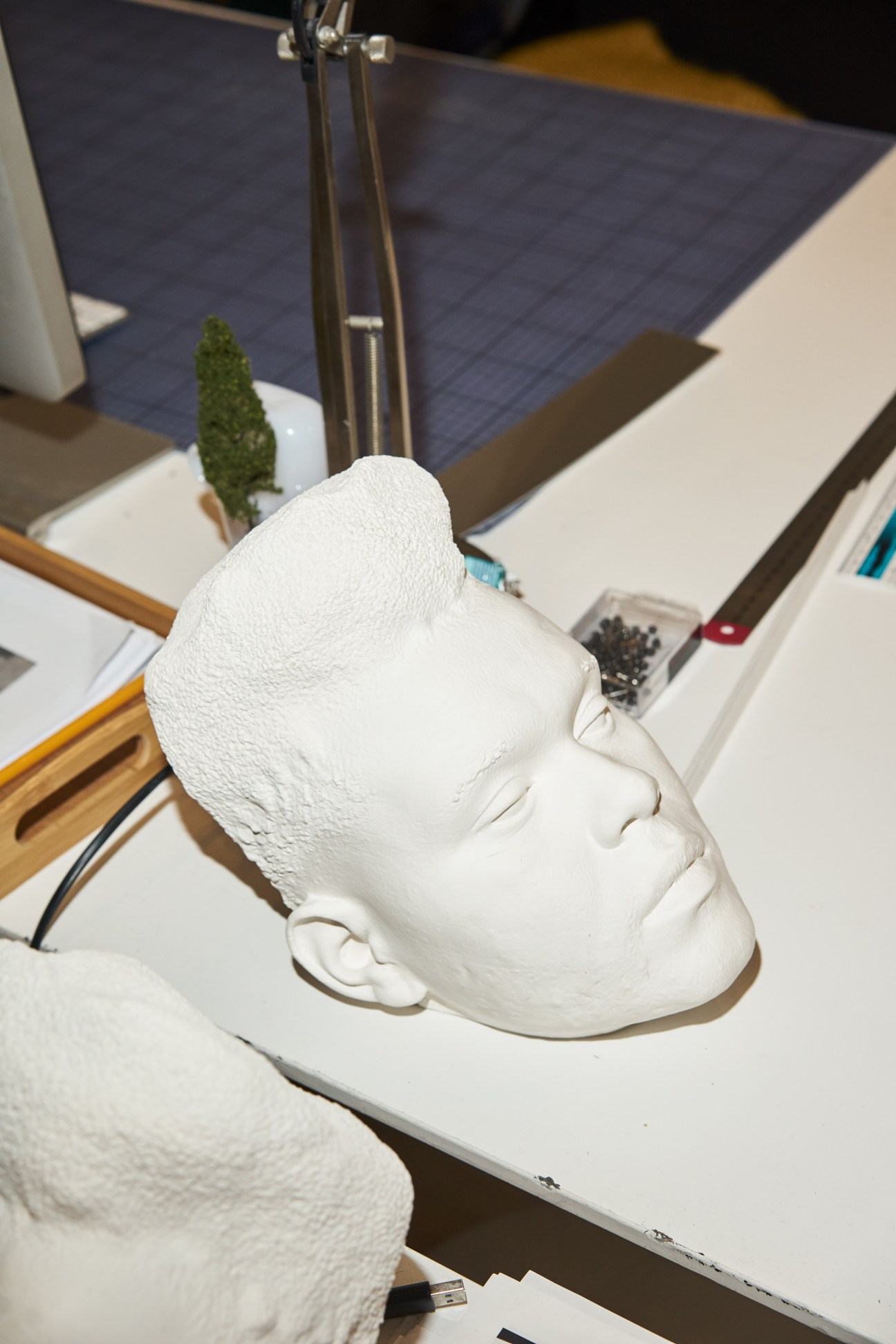 White plaster mask sculpture on a designer's desk with studio materials visible in background.