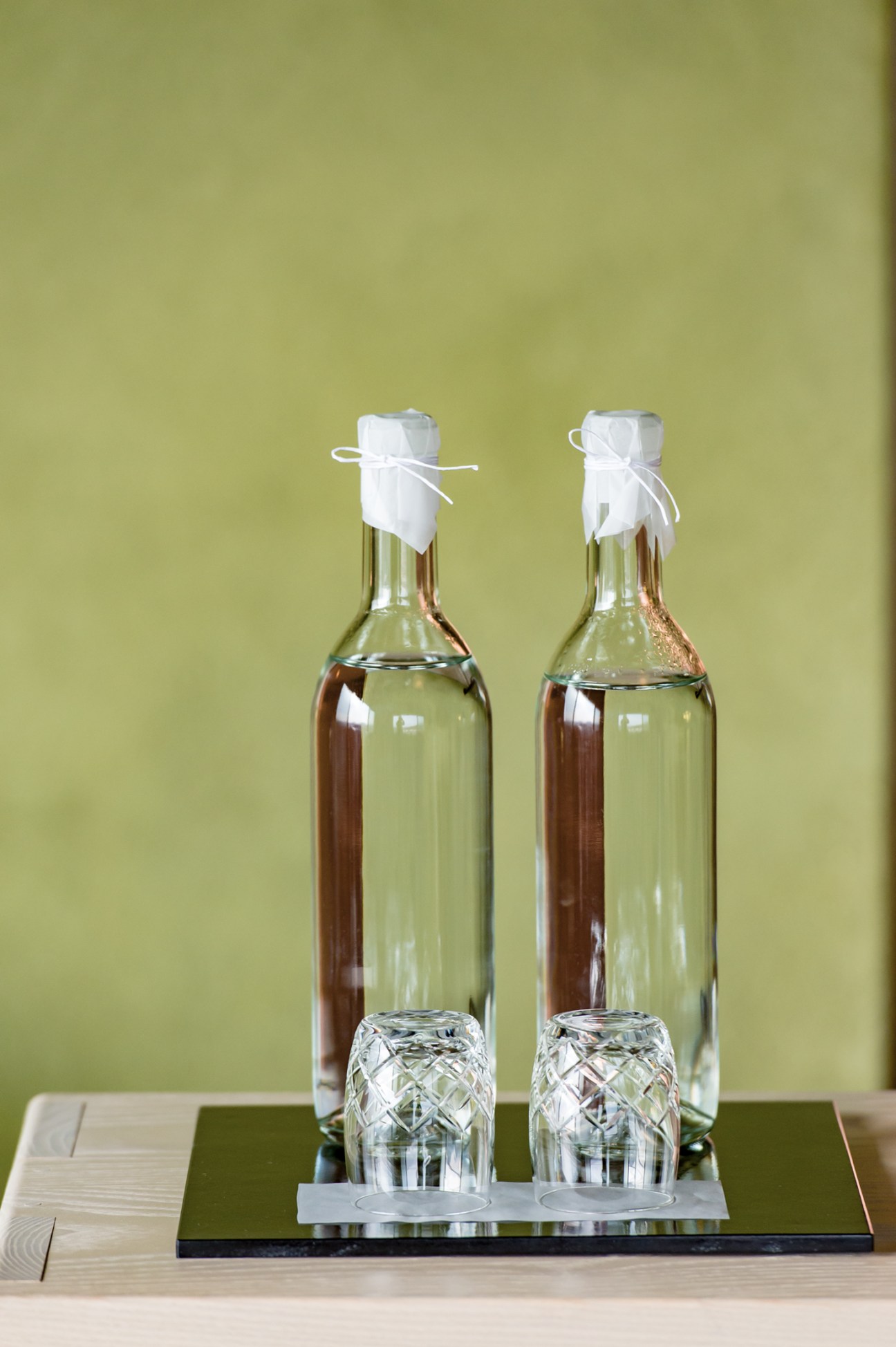 Two glass bottles with white cloth ties and matching crystal glasses on a green surface against a yellow-green wall.