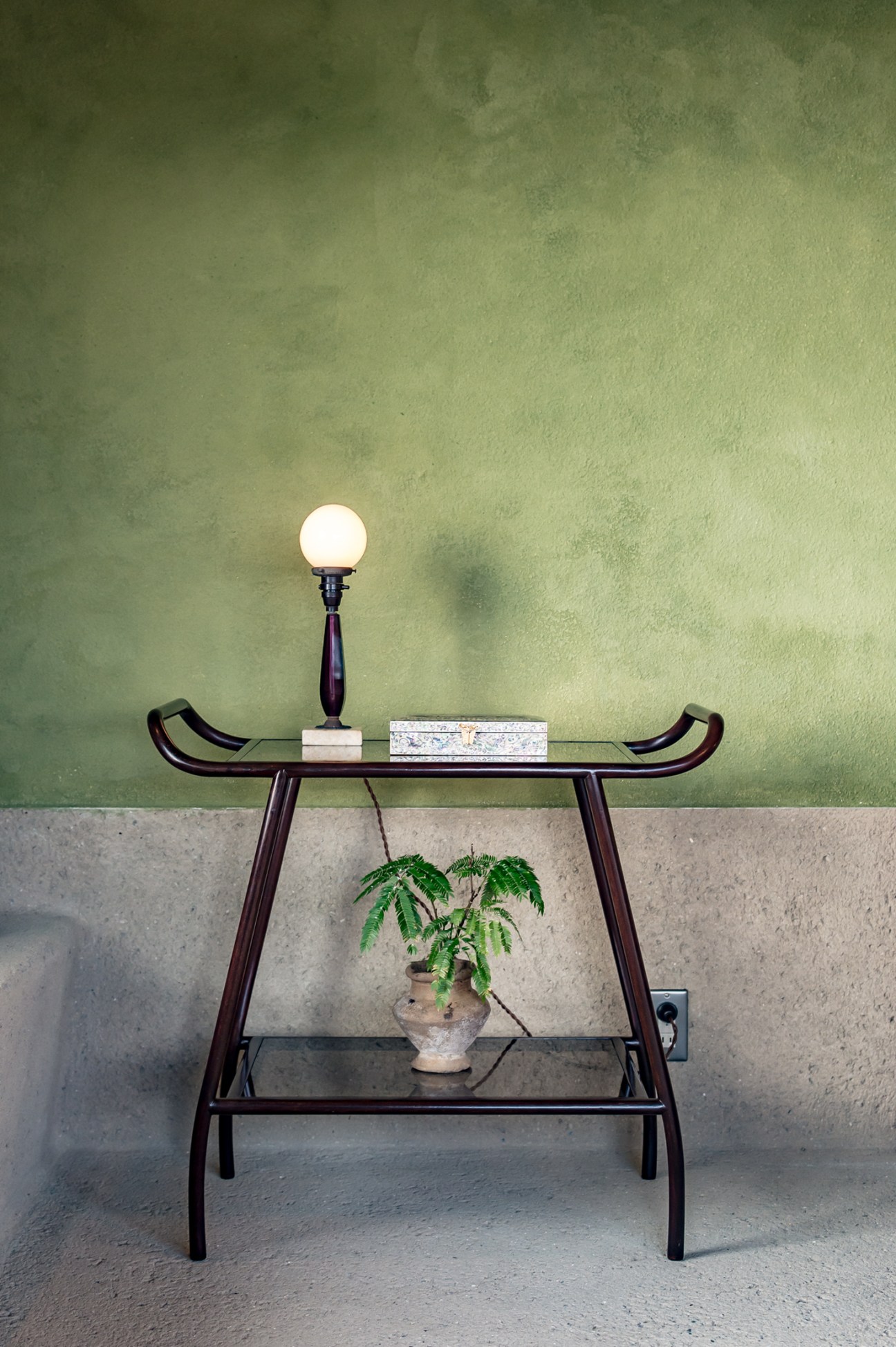 A dark wooden two-tiered table with curved handles against a green textured wall, topped with a glowing globe lamp, books, and a potted plant.