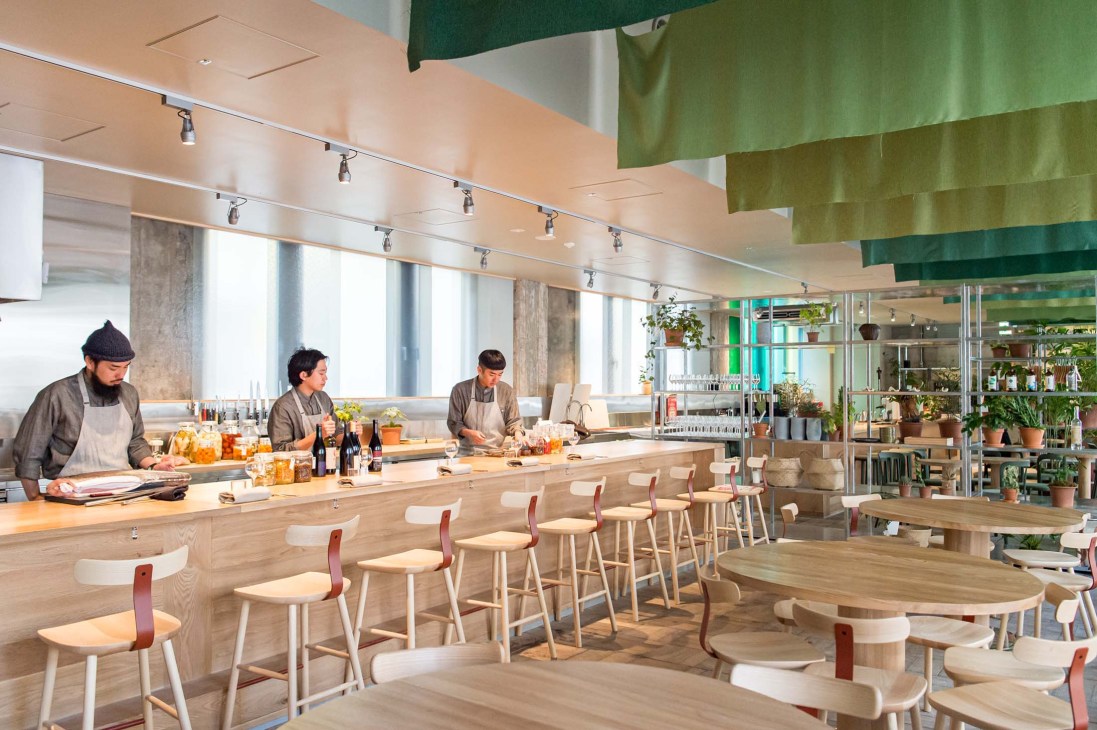 Open kitchen at Caveman restaurant with three chefs working behind counter, wooden bar seating and green ceiling design
