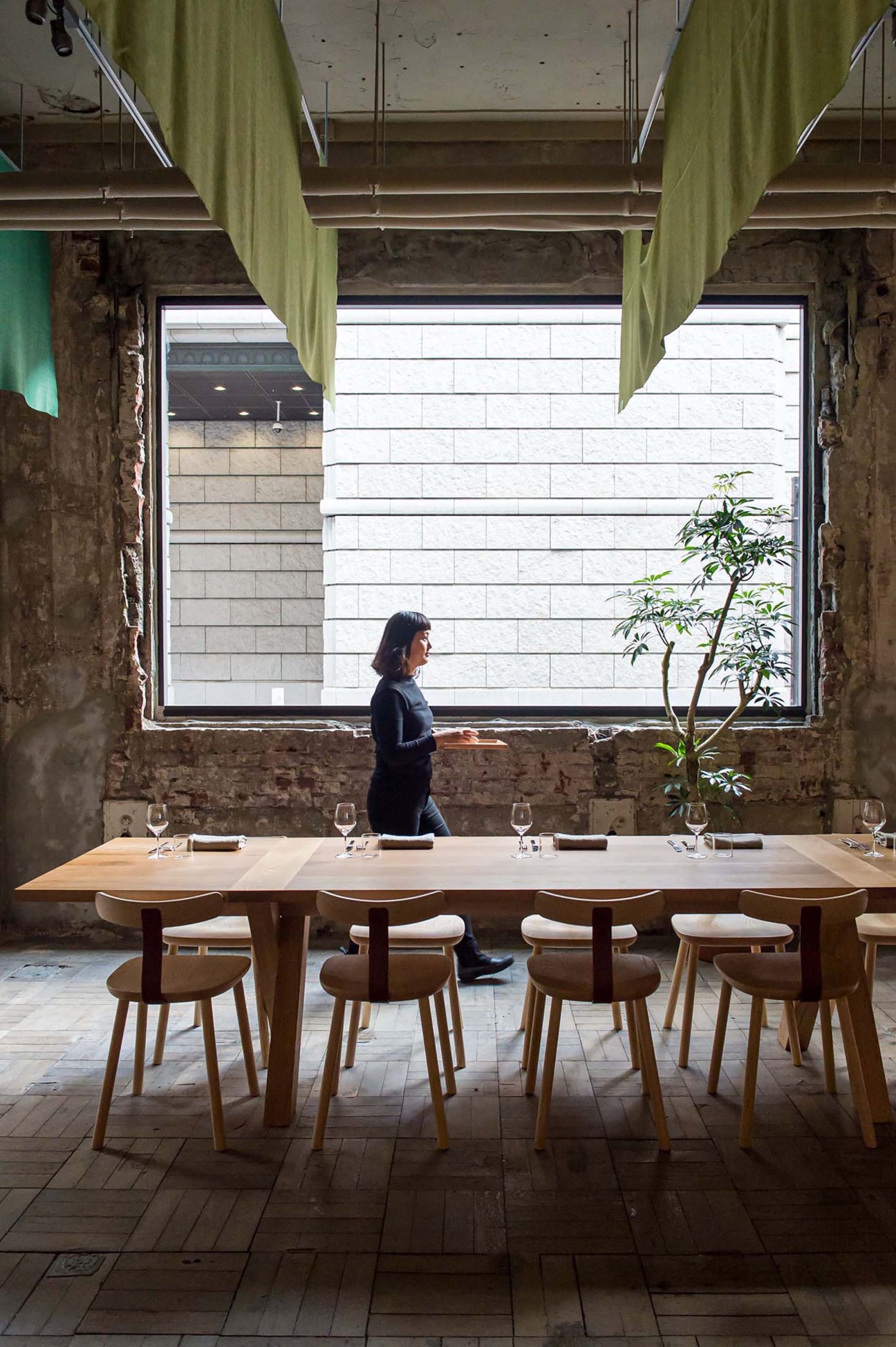 Restaurant interior with long oak dining table, wooden chairs, exposed brick walls, and server walking past large windows