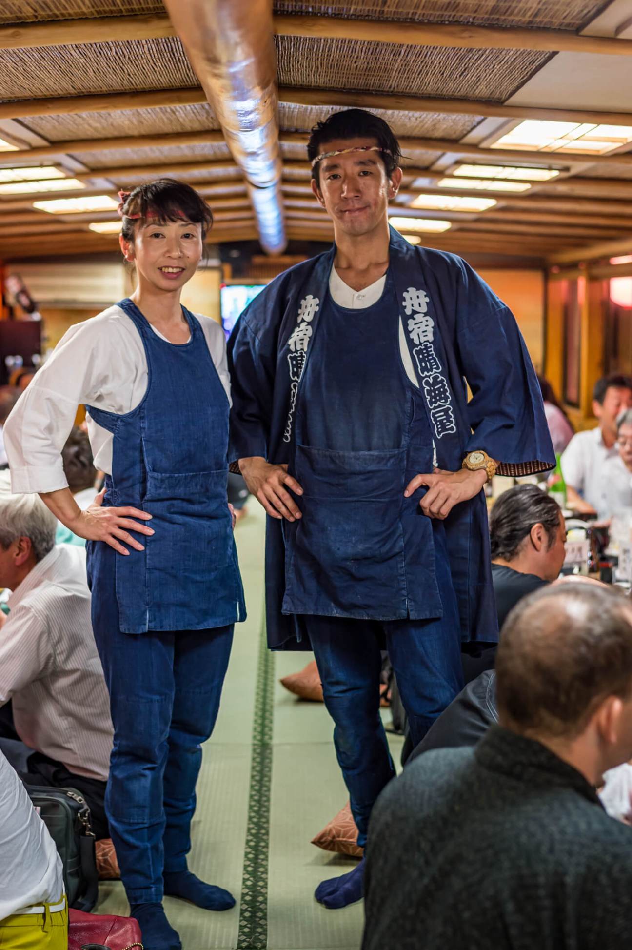 Two staff members in traditional indigo denim uniforms smile aboard a yakatabune river cruise boat.