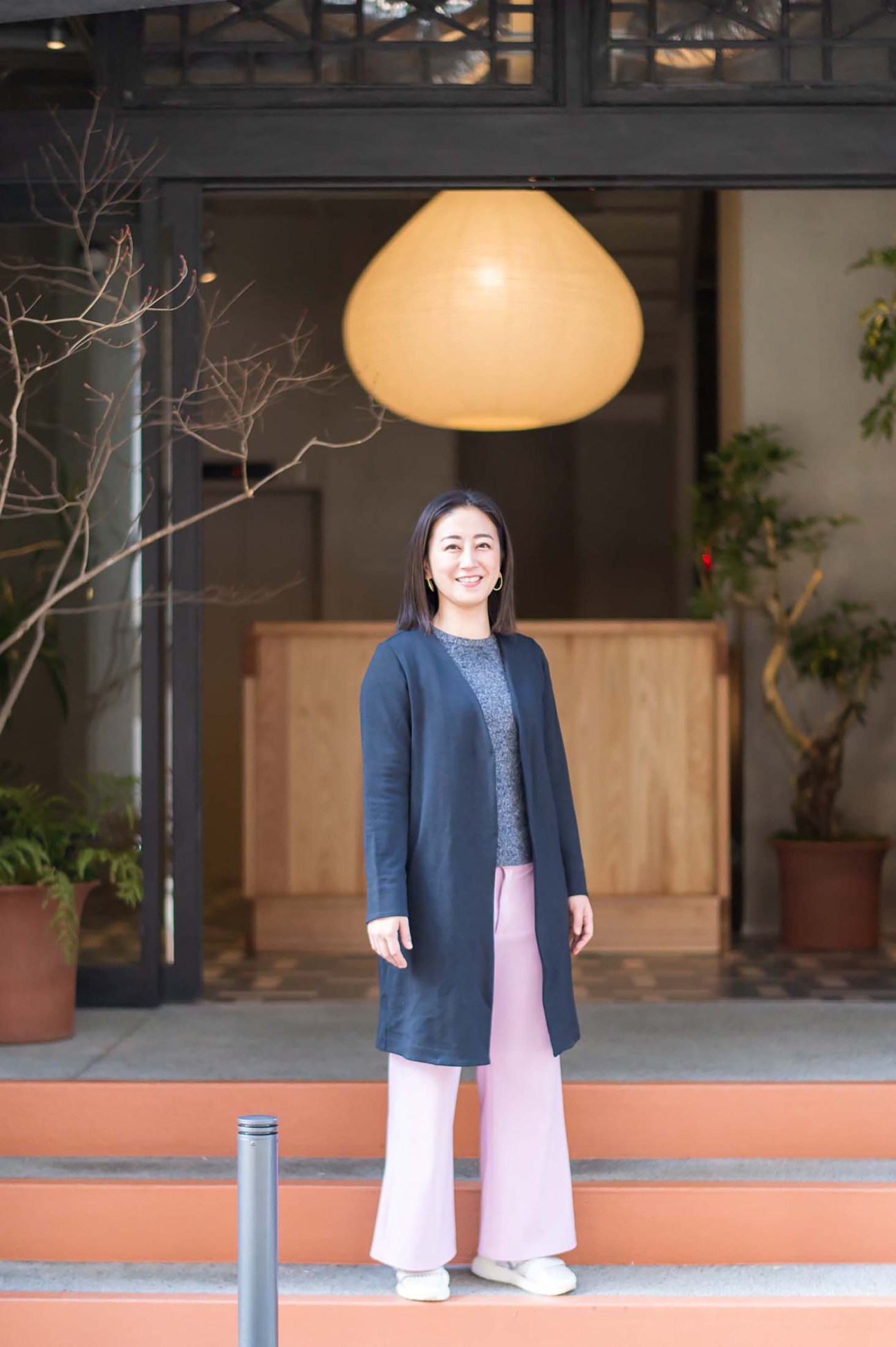 Asian woman in navy coat and pink pants standing outside modern building with large pendant light