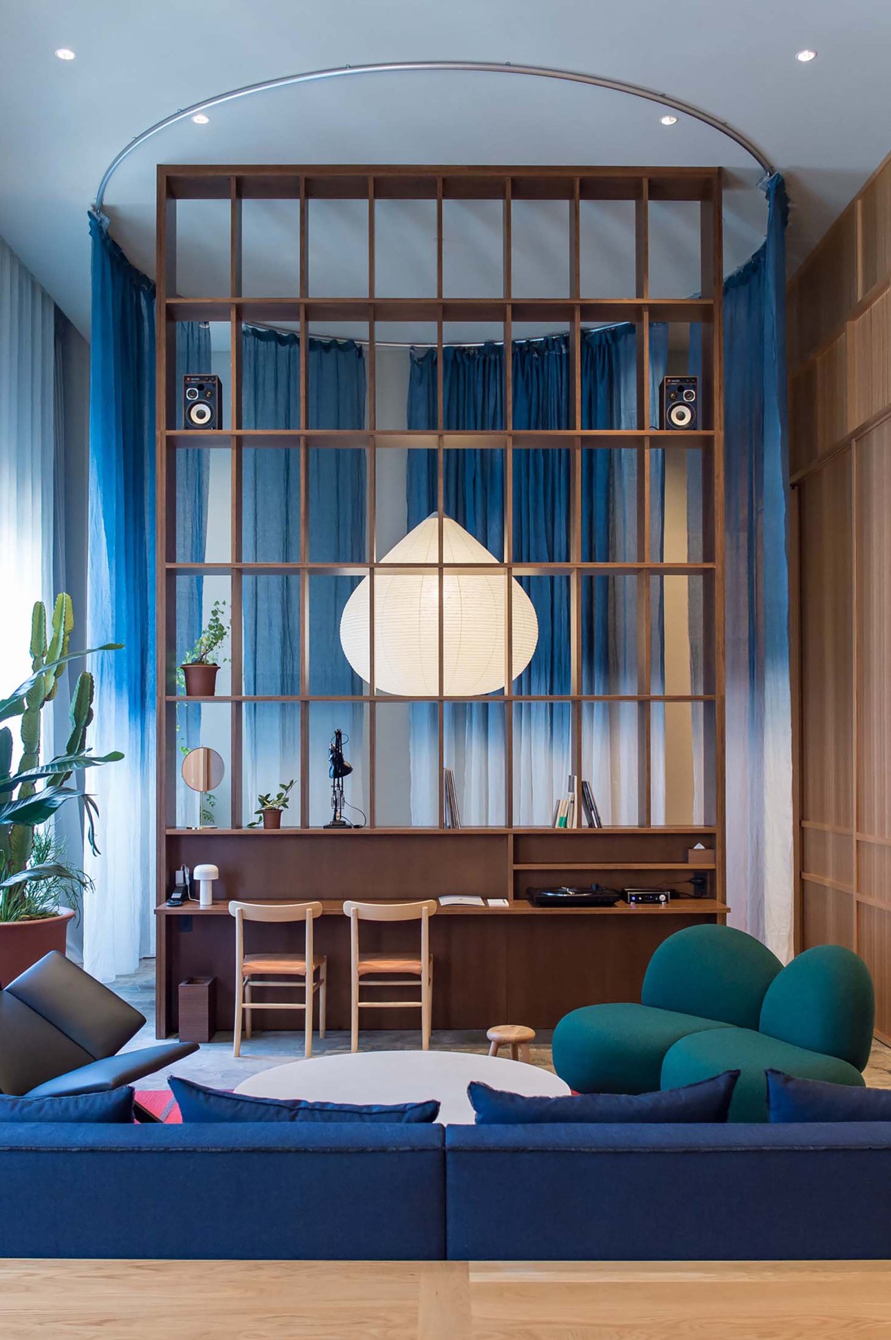Modern hotel room interior with blue sofa, green armchair, wooden shelving unit and circular pendant light