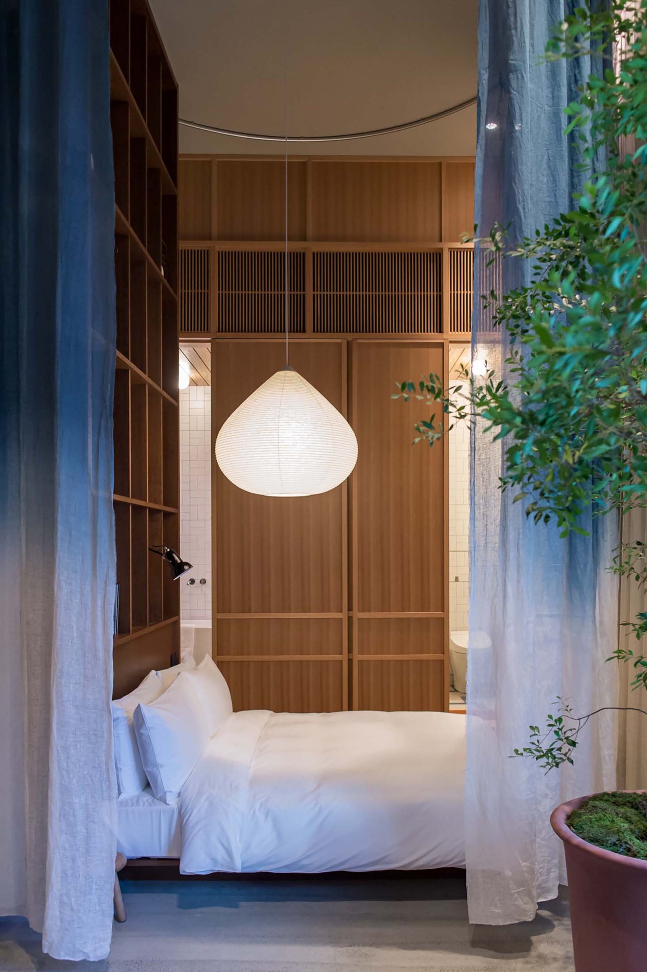 Modern hotel bedroom with white bedding, wooden built-in storage, pendant light, sheer curtains and potted plants