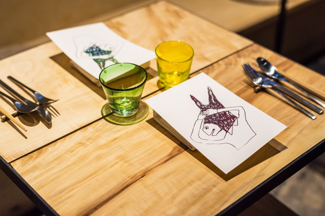 Wooden table setting with ceramic plates, green glasses, cutlery and a sketch drawing at The Blind Donkey restaurant.