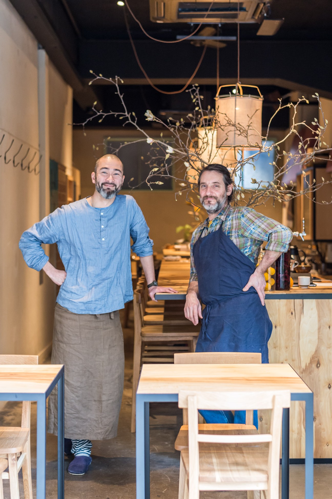 Two men standing in a minimalist restaurant interior with wooden tables and decorative branches hanging from the ceiling.