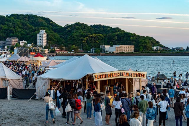 Outdoor beach festival with white tents and crowds gathered around food stalls at sunset near water and forested hills