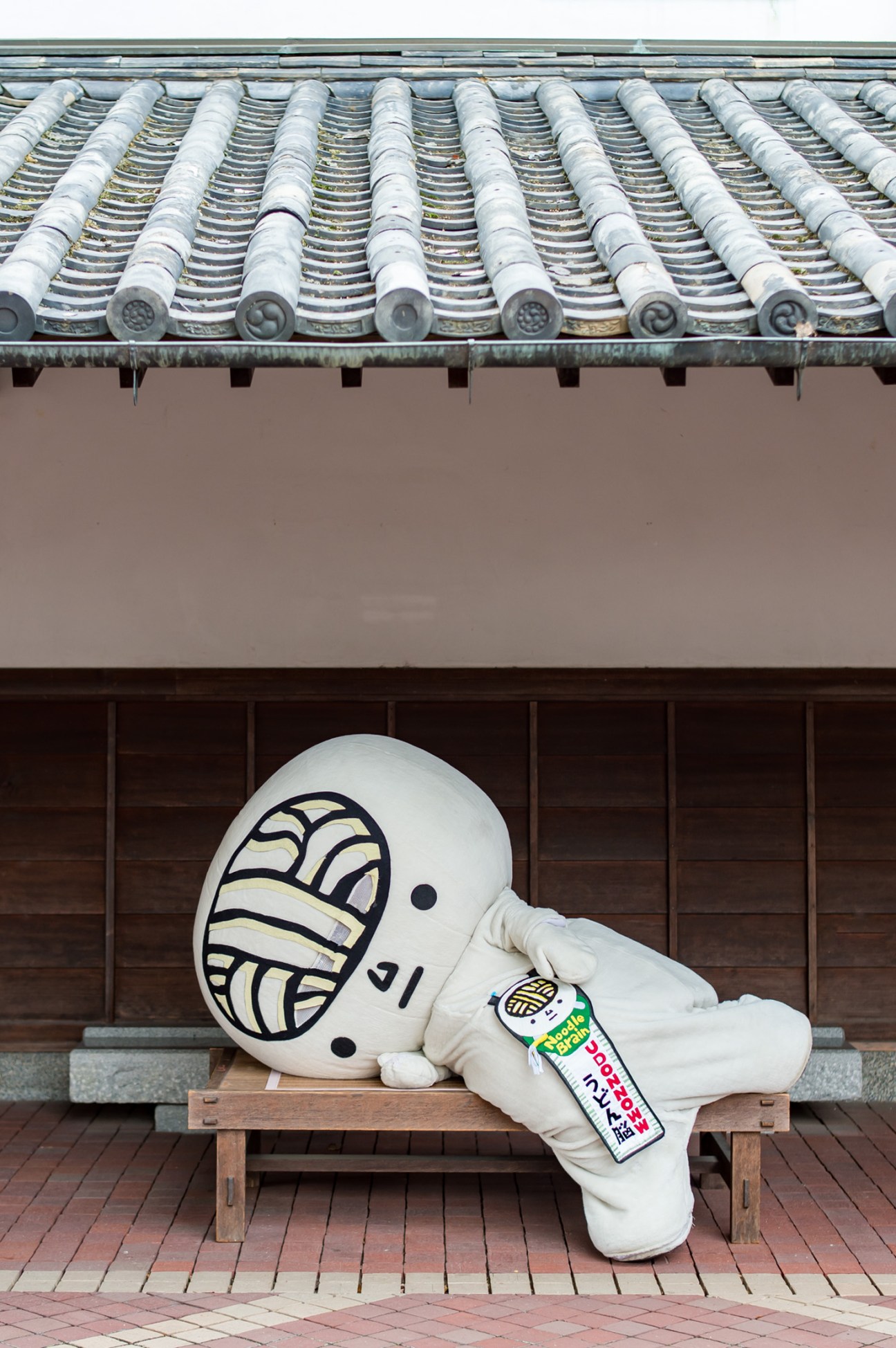 Udon-no mascot character in white costume with noodle design lying on wooden bench