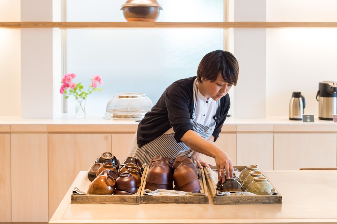 Woman in apron arranging clay bowls and serving vessels on a wooden counter in a bright kitchen.