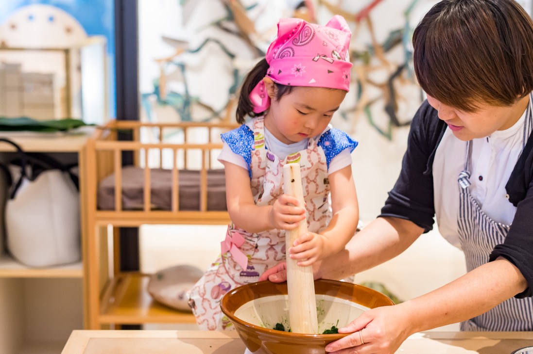 A young girl in a pink headband and apron pounds rice with a wooden pestle while an adult assists at Kageoka no Ie community centre.
