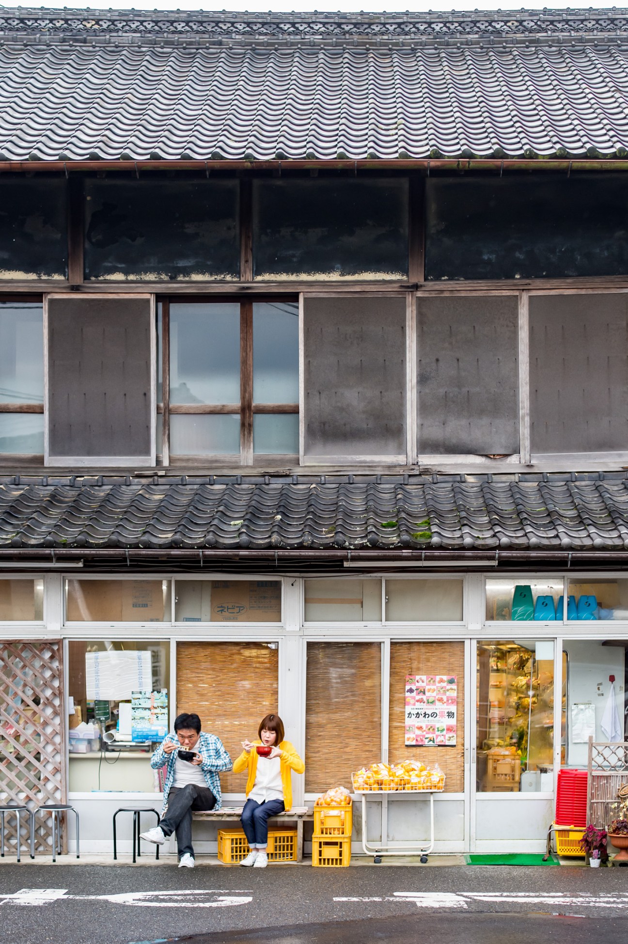 Two people sitting on yellow crates eating outside Suzaki Shokuryohinten grocery shop with traditional tile roof