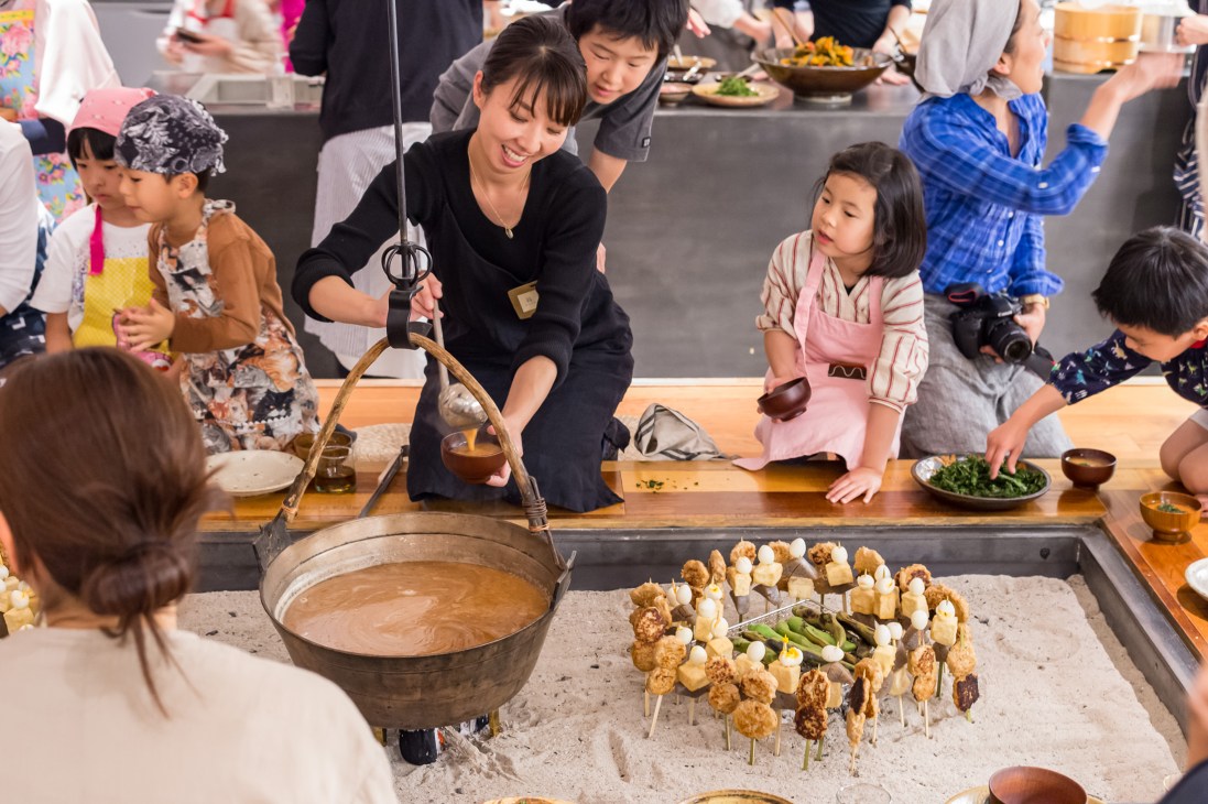 Children and adults cooking tsukune chicken meatballs on skewers over a traditional Japanese irori hearth at Kageoka no Ie community centre.
